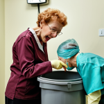 An elderly woman in a dental office, wearing rubber gloves and comically searching through a trash can, looking for her dentures.